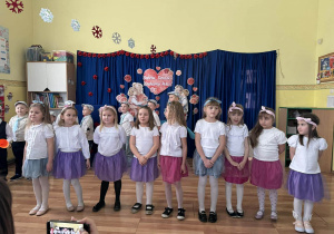 Dziewczynki stoją w rzędzie, ubrane w białe koszulki i spódniczki w kolorach fioletowym, różowym i niebieskim.