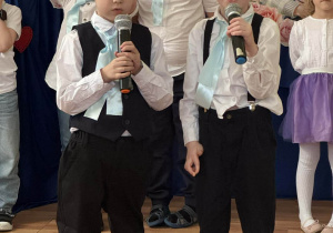 Dwóch chłopców w białych koszulach, jasnoniebieskie krawaty, kraciaste kaszkiety trzyma mikrofony i patrzy w stronę widowni.