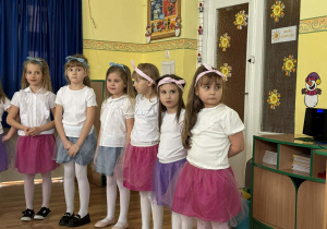 Zdjęcie przedstawia grupę dziewczynek, ustawionych w rzędzie podczas występu. Wszystkie ubrane są w białe bluzki i spódniczki z tiulu w kolorach niebieski, różowy i fioletowy. Mają także białe rajstopy i opaski we włosach w kolorach pasujących do spódniczek. Dziewczynki wyglądają na skupione i gotowe do występu.