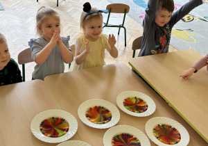 Zdjęcie przedstawia czworo dzieci. Dzieci są podekscytowane i zadowolone ponieważ udał się eksperyment polegający na wykonaniu tęczy z cukierów Skittles. Barwnik rozpuścił się pod wpływem wody i utworzył kolorowe wzory. Przed dziećmi znajduje się biały talerz z kolorowym, przypominającym tęczę wzorem, utworzonym z rozpuszczających się cukierków Skittles w wodzie.