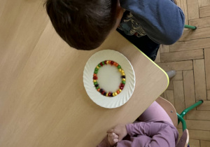 Po prawej stronie znajduje się dziewczynka w fioletowej bluzce, uśmiechnięta i patrząca w obiektyw. Po lewej stronie widoczny jest chłopiec w niebieskiej bluzce, który zasłania twarz rękami. Na stole stoi biały talerzyk. Na talerzyku ułożone są kolorowe draże owocowe Skittles w kształcie okręgu.