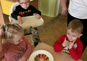 Zdjęcie przedstawia dziewczynkę i dwóch chłopców, obok stoi osoba dorosła. Dwoje dzieci siedzi przy stole, jeden chłopczyk obok w dziecięcym krzesełku. Dzieci są skupione i skoncentrowane. Przed dziećmi znajduje się biały talerz z kolorowym, przypominającym tęczę wzorem, utworzonym z rozpuszczających się cukierków Skittles w wodzie.