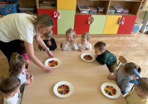 Zdjęcie przedstawia grupę dzieci. siedzących przy stole. Przed dziećmi znajdują się białe talerz z kolorowym, przypominającym tęczę wzorem, utworzonym z rozpuszczających się cukierków Skittles w wodzie.