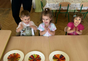 Zdjęcie przedstawia chłopczyka i dwie dziewczynki. Dzieci są uśmiechnięte siedzą przy stole, w sali przedszkolnej. Dzieci są podekscytowane i klaszczą w dłonie. Przed dziećmi znajduje się biały talerz z kolorowym, przypominającym tęczę wzorem, utworzonym z rozpuszczających się cukierków Skittles w wodzie.