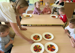 Zdjęcie przedstawia nauczycielkę, która ustywia białe talerze na środku złączonych stołów, na których są wzory utworzone z rozpuszczających się cukierków Skittles w wodzie.