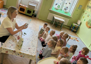 Zdjęcie przedstawia grupę dzieci siedzących na dywanie, przed nimi jest nauczycielka siedząca przy stoliku, która trzyma w ręku butelkę plastikową. opowiada dzieciom o eksperymencie, który przeprowadzi z użyciem octu, balona oraz sody oczyszczonej.