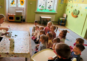 Zdjęcie przedstawia grupę dzieci siedzących na dywanie, przed nimi jest nauczycielka siedząca przy stoliku, która wlewa do pustej butelki ocet.