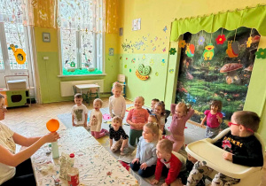 Zdjęcie przedstawia grupę dzieci siedzących na dywanie, przed nimi jest nauczycielka, która nałożyła pomarańczowy balon na butelkę, do którego wczesniej nasypała sodę oczyszczoną. Po nałożeniu na butelkę balonu, wsypała do octu sodę oczyszczoną. W wyniku tego powstał gaz napełniając balon. Dzieci były zaciekawione i szczęśliwe, że eksperyment się udał.