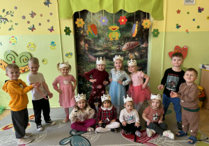 Zdjęcie przedstawia grupę dzieci. Dziewczynki podpierają się w boczki i mają na głowach korony. Chłopcy stoją obok i mają skierowane ręce w kierunku dziewczynek.