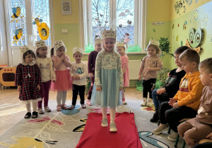 Zdjęcie przedstawia grupę dzieci biorących udział w zabawach z okazji Dnia Kobiet. Dziewczynki mają na głowach korony. Jedna dziewczynka w jasnoniebieskiej sukience stoi na czerwonym dywanie, reszta dziewczynek stoi za nią. Chłopcy siedzą na krzesełkach i biją brawo.