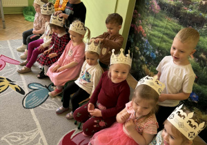 Zdjęcie przedstawia grupę dzieci. Dziewczynki siedzą na krzesełkach, a chłopcy stoją za nimi i próbują wykonać dziewczynkom fryzury.