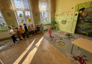 Na dywanie jest tor przeszkód, który składa się z obręczy, stolika, szarfy oraz kolorowych piłeczek. Jedna z dziewczynek skacze do obręczy a reszta grupy się temu przygląda.