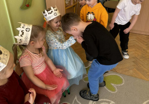 Na zdjęciu widać grupę dzieci bawiących się. Dziewczynki noszą papierowe korony. Centralnym punktem jest chłopiec, który kłania się i całuje w rękę siedzącą dziewczynkę w niebieskiej sukience. Inne dzieci przyglądają się scenie. Atmosfera jest radosna i beztroska.