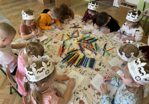 Zdjęcie przedstawia grupę dzieci siedzących wokół stołu i rysujących. Dzieci skupiają się na swoich pracach plastycznych flamastrów rozsypanych na stole.