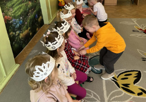 Na zdjęciu widać grupę dzieci bawiących się. Dziewczynki siedzą na krzesłach. Przed nimi stoją chłopcy, którzy się im kłaniają i całują je w rączkę.