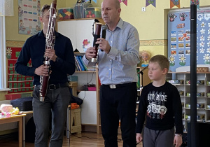 Prowadzący koncert Pan Maciek trzyma w ręce element fagotu. Po jego lewej stronie stoi muzyk trzymający fagot, po prawej stronie chłopiec słucha opisu instrumentu.