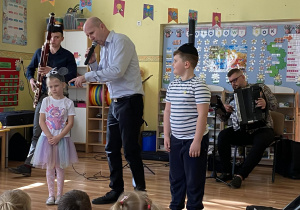 Pośrodku zdjęcia prowadzący śpiewa Balladę o myszy, kocie i fagocie. Po lewej stronie widzimy dziewczynkę z czapeczką myszki na głowie, a po prawej chłopca z czapką kota. Za nimi jeden muzyk gra na fagocie, a drugi na harmonii.