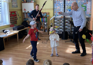 Chłopiec w czapce przedstawiającej bociana idzie w kierunku dziewczynki w pięknym kapeluszu Pani wiosny. Pan Maciek wskazuje palcem na przedszkolaki. W tle widzimy mężczyznę grającego na fagocie.