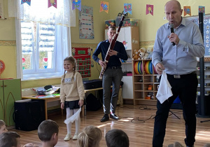 Przy dźwiękach menueta prowadzący, trzymając białą chusteczkę w ręce, idzie w kierunku siedzących przed nim dzieci. Po jego lewej stronie widzimy pana grającego na fagocie i dziewczynkę z białą chustą w ręku. Dziewczynka patrzy na widownię.