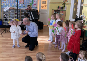 Pan Maciek trzyma za rękę dziewczynkę. Na jej głowie widzimy ogromny kapelusz Pani Wiosny. Po prawej stronie grupka dzieci trzyma w rękach kwiatki. Z tyłu muzyk gra na harmonii utwór pt. Cztery pory roku Viavaldiego - wiosna.