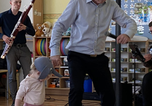 Prowadzący koncert prowadzi za rękę małą dziewczynkę. Na jej główce widzimy czapeczkę przedstawiającą słonika. Po lewej stronie zdjęcia mężczyzna gra na fagocie.