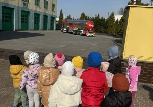 Zdjęcie przedstawia grupę dzieci patrzących patrzących na wjeżdżający na plac czerwony wóz strażacki.