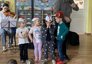 Trzy dziewczynki i chłopiec słuchają utworu Mozarta Kurki Trzy. Na głowach dziewczynek widzimy czapeczki kur, a na głowie chłopce czapkę koguta. W tle pani gra na skrzypcach.