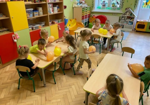 Dzieci siedzą przy stolikach i dmuchają na leżące przed nimi balony.