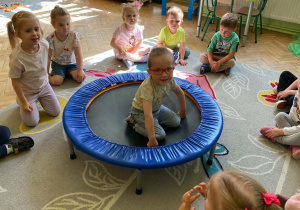Chłopiec siedzi na trampolinie. Dookoła widzimy grupę dzieci siedzących na dywanie.