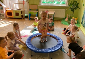 Dziewczynka skacze na trampolinie. Wokół pozostałe dzieci siedzą na dywanie i czekają na swoją kolej.