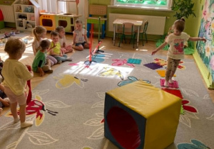 Dziewczynka pokonuje tor przeszkód - przechodzi po specjalnych klockach w kształcie kwadratów. Po lewej stronie pozostałe dzieci siedzą na brzegu dywanu dopingując koleżankę.