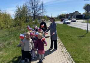 Dzieci z biało-czerwonymi flagami spacerują chodnikiem wzdłuż ulicy.