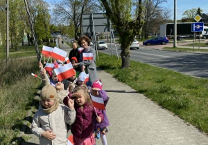 Przedszkolaki niosą flagi, maszerując chodnikiem.
