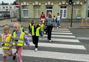 Dzieci wraz z panią przechodzą przez przejście z sygnalizacją świetlną. W tle sygnalizator z zielonym switłem.