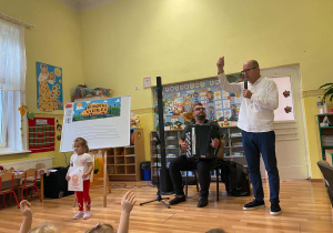 Pan Maciek trzyma mikrofon w dłoni, Pan Michał siedzi z akordeonem i na nim gra, dziewczynka trzyma rysunek nutki o imieniu Celinka.