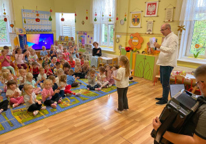 Dzieci aktywnie uczestniczą w koncercie klaszczą w ręce według podanego rytmu przez prowadzącego. Przed dziećmi stoi dziewczynka