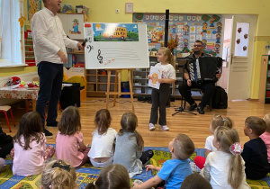 Prowadzący rozmawia z dziewczynką przy tablicy, na której widzimy narysowaną pięciolinię, na niej klucz wiolinowy i przypięte nutki. Dziewczynka trzyma w rękach rysunek nutki o imieniu Ewa.