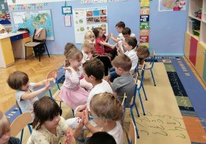 Dzieci siedzą naprzeciwko siebie na krzesełkach i wykonują rytmiczne zabawy przy muzyce.