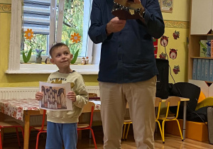 Mężczyzna w okularach i granatowej koszuli trzyma w rękach mały miech oraz mikrofon, a stojący obok niego chłopiec prezentuje rysunek przedstawiający organy kościelne.