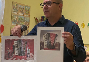 Mężczyzna w okularach i granatowej koszuli uśmiecha się, trzymając w rękach mikrofon oraz dwie ilustracje, z których jedna przedstawia wpisany do Księgi Rekordów Guinessa największy akordeon na świecie.