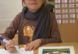 Dziewczynka w kraciastej czapce, dużych okularach i szarym szaliku siedzi przy stole, trzyma marker nad kartką papieru, a obok leży odwrócone zdjęcie wieży Eiffla.